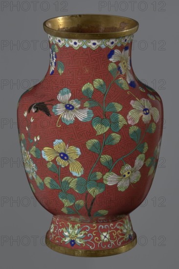Vase, gift; Chinese, Qing dynasty Xianfeng, 1851–1861; cloisonné enamel. The vase is set on a tall, conical foot flaring downwards, terminating in a metal ring, decorated with a lotus tendril. The short, wide neck is topped with a metal ring below which is a border composed of ruyi heads. The belly and neck are decorated with an all-around composition of flowers and plum branches against a Chinese meander background on a red ground. 1st half of the 19th century; vessel. Height 25.1 cm; diameter 16.5 cm; weight 1.5 kg., A rounded decorative vase photographed against a neutral gray background, predominantly red with a repeating textured pattern across its body, featuring floral motifs with a few flowers in white, pale yellow, light blue, and pale pink, each with dark blue central accents, and green leaves and stems. The rim and base have metallic gold bands, and the base area includes ornate scrolling and floral designs in pale green, white, blue, and gold. There is also a small dark fea...