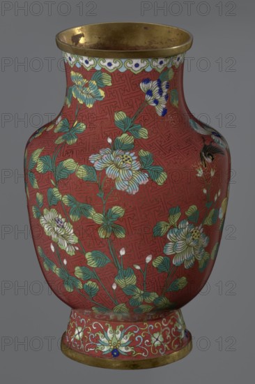 Vase, noted; Chinese art; Qing dynasty 1644–1911, Xianfeng period 1851–1861; cloisonné enamel; floral motifs and meander ornament. The vase is set on a tall, conical foot flaring downwards and terminating in a metal ring, decorated with a lotus tendril. The neck is short and wide, also topped with a metal ring, below which is a border composed of ruyi heads. The belly and neck are decorated with a continuous composition of flowers and plum branches against a red Chinese meander background. 1st half of the 19th century; China; vessel. Height 25.1 cm, diameter 16.5 cm, weight 1.5 kg., A rounded vase with a flared rim and a slightly footed base sits against a neutral gray background; the vase surface is predominantly red with an allover geometric pattern and is decorated with a group of stylized flowers and leaves in shades of green, teal, pale yellow, white, and light blue, with some flower accents in dark blue and black; the rim and base edges are metallic gold and show some darker spot...