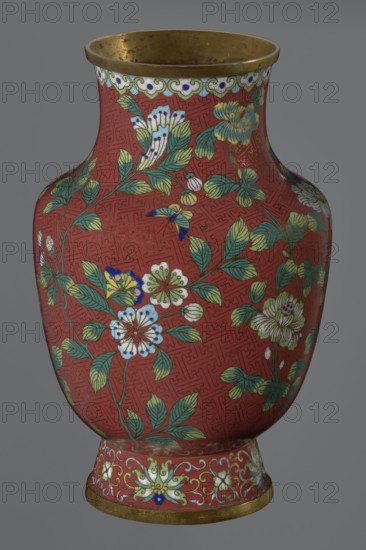 Vase, gift; Chinese art; Chinese; Qing dynasty 1644–1911, Xianfeng period 1851–1861; cloisonné enamel; floral and meander ornamentation. The vase stands on a tall, conical foot flaring downward to a metal ring, decorated with a lotus tendril. The short, wide neck is topped with a metal ring and a border of ruyi-heads. The belly and neck are covered all around with a composition of flowers and plum branches against a red Chinese meander background. 1st half of the 19th century. Vessel; height 25.1 cm, diameter 16.5 cm, weight 1.5 kg., A rounded vase with a narrow neck and flared rim sits against a plain gray background; the vase is predominantly red with a repeating maze-like textured pattern and is decorated with a floral design featuring green leaves and flowers in white, yellow, blue, and pale green, along with small accents of dark blue and gold; the rim and base have a metallic gold band, and the base area shows an additional floral border in pale green, white, and blue on the red ...