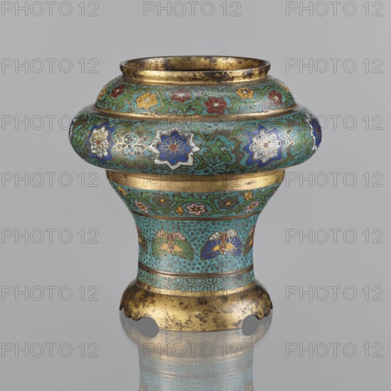 Vessel, Japanese, Meiji period 1868–1912, 19th century. Materials/technique: champlevé enamel. Decoration: plant motifs, butterflies, flowers, fans; sasarindo coat of arms. Dimensions: height 28 cm; diameter 28.6 cm., An ornate vessel with a rounded upper section narrowing into a flared base sits against a plain light gray background; its surface shows a turquoise green patina with intricate floral and vine motifs in shades of blue, white, red, yellow, pink, and green, accented with metallic gold bands and gilt areas that show darker wear; the upper rim and base edges are gold-toned with irregular dark spots, and the piece displays repeating stylized flower medallions and leaf-like patterns around the midsection, with a reflective surface beneath suggesting a faint reflection.