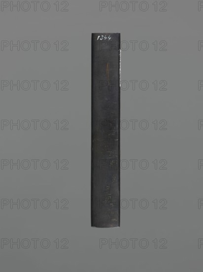 Knife handle kozuka, gift; Japanese, Edo–Meiji period 19th century, 1603–1912; weapons/edged weapon for a katana kozuka; materials not specified; height 9.8 cm, width 1.5 cm., A vertical rectangular object with a worn dark surface centered against a uniform gray background, showing a few scratches and scuffs and a thin lighter streak along one edge, with visible subtle variations of dark gray and black tones and a small patch of brownish-orange discoloration.