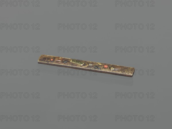 Knife handle kozuka, Japanese, Edo–Meiji period 19th century, Edo/Meiji, 1603–1912, weapons/edged weapon for katana, kozuka; materials unspecified; height 9.8 cm, width 1.5 cm., A long narrow rectangular object with a metallic, worn surface in brown and gold tones rests on a smooth grey background; the object bears small decorative insets in red and green and darker accents, and shows mottled gold flecks and patches along its length, with a subtle reflection visible on the background.
