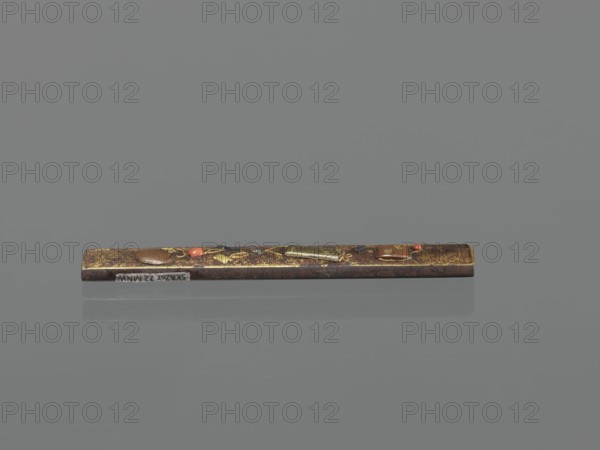 Knife handle kozuka, Japanese, Edo–Meiji period 19th century, Edo/Meiji, Japan. Materials/subject: weapon accessory for katana kozuka, inrô, netsuke context; oriental edged weapons. Dimensions: height 9.8 cm, width 1.5 cm., A narrow rectangular object rests horizontally against a smooth background, showing worn brown and gold surfaces with small raised decorative elements in metallic gold and darker brown, along with a few small accents of red and black; the background and surface reflection are shades of gray.