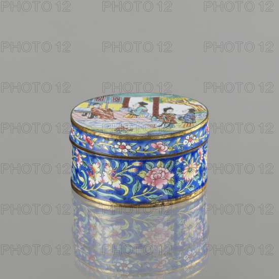 Box with a lid;, painted enamel; Qing dynasty 1644–1911; 19th century; Vietnamese Nguyen dynasty, 1802–1945 and Chinese cultural influence; figurative scenes depicting peonies, grapes, and peaches iconography; container; height 3.3 cm, diameter 6.3 cm., A small round box with a domed lid sits on a reflective surface, showing a bright palette of blue, pink, green, yellow, white, gold, and brown. The box exterior is predominantly cobalt blue with painted floral motifs in pink, white, and yellow with green leaves and gold accents that form a decorative band around the sides. The lid features a painted scene with small figures and architectural elements rendered in muted tones of yellow, brown, blue, green, and pink, bordered by a thin gold rim. The reflection of the box on the surface repeats the blue and floral patterns and soft colors of the lid.