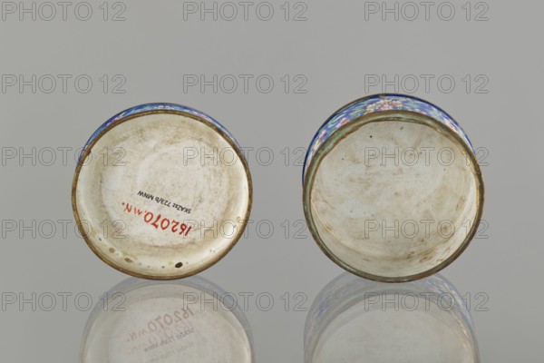 Box with lid; creator, painted enamel; 19th century, Qing 1644–1911 and Nguyen dynasty 1802–1945 — China/Vietnam place of origin uncertain; figurative scenes of peonies, grapes, and peaches; Vietnamese and Chinese art influences; container; height 3.3 cm, diameter 6.3 cm., Two round shallow bowls shown from above resting on a reflective surface, each with an off-white interior that has visible smudges and worn marks; the rims are edged with a metallic band and decorated exterior patterns in blue with accents of pink, green, and white; the left bowl shows some small dark spots and an area of reddish marks on its inner surface, while the right bowl has a more uniformly stained interior with subtle brownish streaks; the reflection beneath them mirrors their shapes and colors against a neutral gray background.
