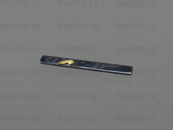 Knife handle kozuka, gift; Japanese; Edo period 1603–1868, 19th century; weapon knife handle for a katana; materials/technique: metal with decorative inlay depicting bats and pines; height 9.7 cm, width 1.5 cm., A long, thin rectangular object rests on a flat surface, appearing glossy and dark with a reflective sheen; the object is primarily black with a metallic luster and has a small raised decorative element in a bright gold color near one end, along with subtle lighter highlights and faint reflections on the surface beneath it; the background and surface are uniform shades of gray.
