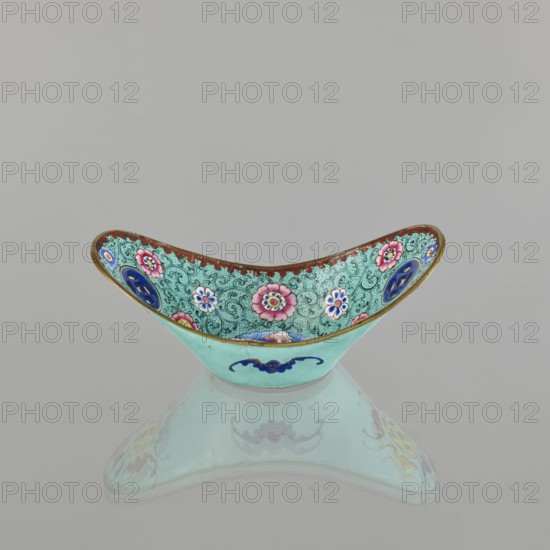 Bowl; painted enamel on porcelain; Qing dynasty 1644–1911, 18th–19th century, China; decorated with lotuses, coins, flower tendrils and bats symbolizing happiness; vessel height 6.3 cm, width 8.5 cm, depth 15.3 cm., A shallow, crescent-shaped decorative bowl sits on a reflective surface, showing a mirrored image beneath. The bowl's exterior is a pale turquoise with a dark blue stylized motif on the front, while the interior displays an intricate patterned background in a slightly darker turquoise filled with swirling designs and floral medallions in pink, white, and blue, each with contrasting centers. The interior rim has a narrow band of rusty red edged with a thin gold border, and there are additional circular blue medallions set into the patterned interior. The overall color palette includes shades of turquoise, pink, blue, white, rusty red, gold, and small accents of darker tones.