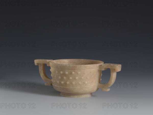 Gui-type bowl, Chinese, Qing dynasty 1644–1911, 2nd half 18th century. Porcelain vessel modeled after a ritual gui. Supported by a low ring foot; walls decorated with three horizontal rows of small convex knobs evoking archaic ruding nailheads, framed by a thin rim at the bottom and a band framing the spout at the top. Two stylized handles on either side reference the ancient prototype. Upper part decorated with an image of cicada larvae arranged in a rosette. Height 4.5 cm; width 13 cm; diameter 7.2 cm., A shallow, round cup-like vessel with a warm beige color sits centered on a smooth surface, featuring a band of raised rounded bumps around its exterior; it has a short pedestal base and two curved handles extending outward on opposite sides that are the same beige tone, and the background transitions from a dark gray at the top to a lighter gray near the surface.