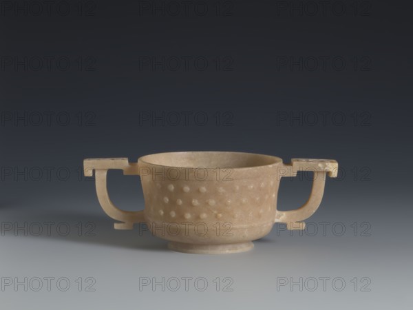 Gui-type bowl, Qing dynasty 1644–1911, 2nd half 18th century. Bronze vessel modeled after a ritual gui. The bowl is supported by a low, ring-shaped foot; its shape and decoration refer to a ritual gui vessel. The walls are decorated with three horizontal rows of small, convex knobs reminiscent of archaic ruding nailheads decoration, framed by a thin rim at the bottom and a band framing the spout at the top. Two stylized handles on either side echo the old prototype. The upper part is decorated with an image of cicada larvae arranged in the form of a rosette. China. Height 4.5 cm; width 13 cm; diameter 7.2 cm., A shallow, round bowl with a short foot and two symmetrical angular handles sits centered on a smooth surface, the bowl carved from a warm beige stone with lighter and darker beige variations; the body of the bowl shows a textured band of raised rounded bumps around its midpoint, and the handles extend outward with right-angled inner and outer profiles. The background is a smooth...