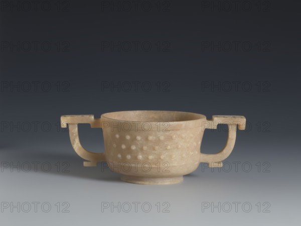 Gui-type bowl, Chinese, Qing dynasty 1644–1911, 2nd half 18th century. Porcelain vessel referencing a ritual gui shape; supported by a low ring-shaped foot. Walls decorated with three horizontal rows of small convex knobs reminiscent of archaic ruding nailhead decoration, framed by a thin rim at the bottom and a band framing the spout at the top. Two stylized handles on either side echo the ancient prototype. Upper part decorated with cicada larvae arranged in a rosette. Height 4.5 cm; width 13 cm; diameter 7.2 cm., A pale beige stone bowl with two symmetrical handles sits centered on a smooth gray surface against a dark gray gradient background; the bowl has a rounded body with a banded base and a pattern of raised rounded bumps on its exterior, and the handles extend outward with rectangular top and bottom elements, all showing subtle variations of beige, cream, and light brown tones.