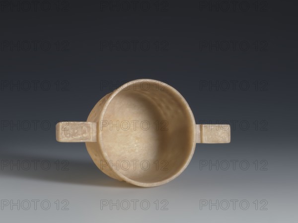 Gui-type bowl, Qing dynasty 1644–1911, 2nd half 18th century. Porcelain vessel referencing a ritual gui. Supported by a low, ring-shaped foot; walls decorated with three horizontal rows of small convex knobs reminiscent of archaic ruding nailheads. Ornament framed by a thin rim at the bottom and a band framing the spout at the top. Two stylized handles on either side echo the old prototype. Upper part decorated with an image of cicada larvae arranged in a rosette. China. Height 4.5 cm; width 13 cm; diameter 7.2 cm., A shallow round vessel with a smooth interior and two short rectangular handles extending from opposite sides, shown from above against a gradient background that transitions from dark gray at the top to lighter gray at the bottom; the vessel and handles are a warm beige to light tan color with subtle variations and a slightly matte surface, and a soft shadow falls beneath and slightly to the left of the vessel.