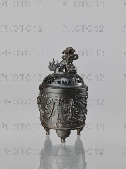 Censer with cover, Chinese, Qing dynasty 1644–1911, 18th century. Material/Type: ritual/ceremonial vessel censer, height 18.5 cm, width 11.2 cm. Decoration/iconography: lions, Immortals, fo, Taoism, lingzhi., A small ornamental vessel stands on a reflective surface against a plain light gray background, featuring a dark metallic color with areas of brownish patina; the vessel has a domed lid topped by a sculpted animal figure with a curled tail and a rounded object beneath one paw, and the lid shows openwork cutouts; the cylindrical body is decorated in low relief with scenes of human figures, trees, and flowing forms, and it rests on three short rounded legs that mirror in the surface below.