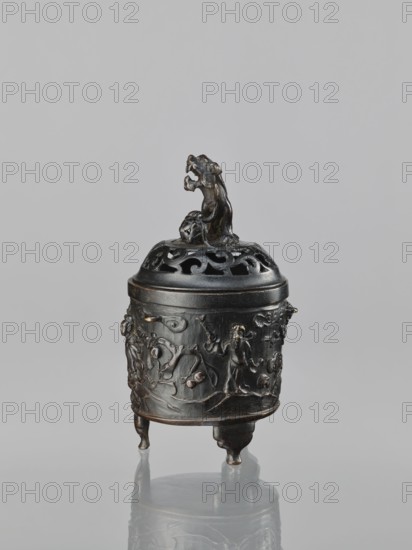 Censer with cover, Chinese, Qing dynasty 1644–1911, 18th century. Ceremonial ritual vessel censer, height 18.5 cm, width 11.2 cm. Decoration: lions, Immortals, fo, lingzhi; Taoist iconography., An ornate dark metal cylindrical container with a removable domed lid topped by a sculpted animal figure, resting on three short legs; the body of the container is decorated with low-relief scenes of human and animal figures and flowing motifs, and the lid has openwork patterns beneath the sculpted topper; colors visible include dark brown, black, and subtle metallic highlights against a pale gray background.