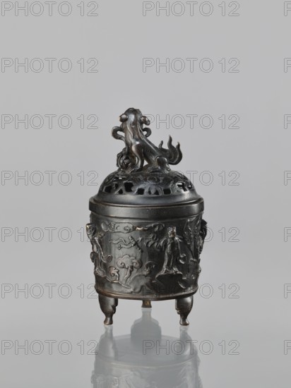 Censer with cover, Qing dynasty 1644–1911, 18th century, China. Ritual/ceremonial vessel censer, height 18.5 cm, width 11.2 cm. Decoration/iconography: lions, Immortals, fo, lingzhi; Taoist associations. Materials and style: Chinese art, Qing style., A dark metallic tripod vessel with a rounded body and a removable domed lid topped by a sculpted animal figure, all in shades of dark brown and black with subtle worn highlights; the body rests on three short legs and is decorated in low relief with carved human figures, stylized clouds, and organic forms; the lid features pierced details and a curled tail on the animal finial; the surface shows areas of patina and lighter brown wear against an overall matte dark finish, set against a plain light gray background with a soft reflection beneath the object.