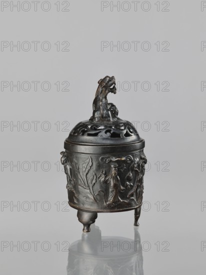 Censer with cover, Qing dynasty 1644–1911, 18th century, China; ritual/ceremonial vessel; materials not specified; decorated with lions, Immortals, fo, lingzhi; Taoist iconography; height 18.5 cm, width 11.2 cm., A small dark metal vessel with a lid stands on three short legs, the lid topped by a sculpted animal figure; the vessel's surface is decorated with raised floral and foliate motifs and stylized figures in low relief. The metal appears dark brown to black with subtle lighter brown highlights on raised areas. The background is a smooth light gray. The vessel casts a faint reflection on the glossy pale gray surface beneath it.