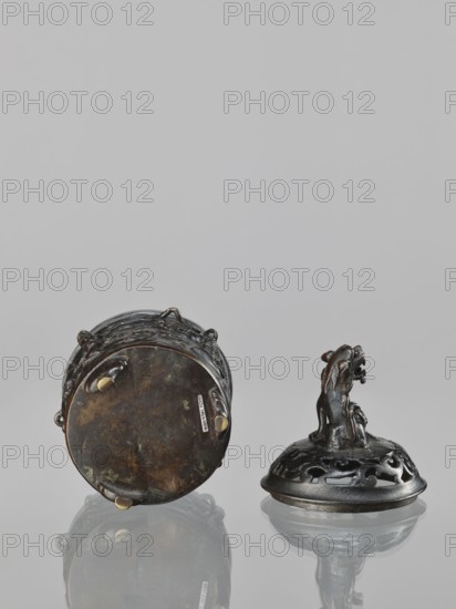 Censer with cover, Chinese, Qing dynasty 1644–1911, 18th century. Ceramic or metal vessel used as a censer/ceremonial ritual vessel; height 18.5 cm, width 11.2 cm. Decoration: lions, Immortals, fo, Taoist imagery, lingzhi., A reflective surface holds a group of two dark metal objects with an aged patina; one object lies on its side showing a round bottom with small rounded feet and irregular surface discoloration in brown and dark tones, while the other stands upright displaying an openwork domed top with flowing cutout patterns and a sculpted animal-like figure rising from the center with a textured body and raised head; the colors visible include dark gray, brown, muted gold highlights on small feet, and subtle greenish oxidation, all set against a pale gray background with faint reflections beneath the objects.