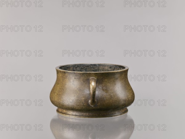 Censer, Chinese, Qing dynasty 1644–1911, 18th century; ceremonial/ritual vessel; backdated signatures; material not specified; height 8.5 cm, diameter 16.8 cm., A low, rounded metal vessel with a bulbous body and a slightly flared rim sits centered on a reflective surface; its exterior shows mottled tones of brown, dark green, and muted gold with a patinated finish, and a small curved handle-like projection is visible on the front, while the interior appears darker with grayish and black hues and the background is a soft pale gray.