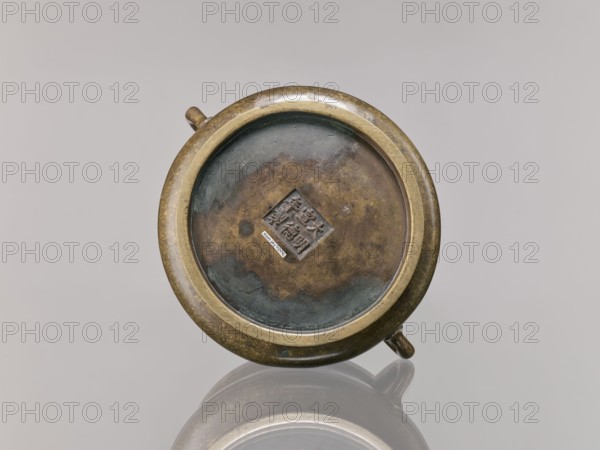 Censer, Chinese, Qing dynasty 1644–1911, 18th century; ceremonial/ritual vessel; backdated signatures; bronze censer; height 8.5 cm, diameter 16.8 cm., A round metal object with a circular rim and a recessed center sits against a plain light gray background, showing a patina of brown, green, and dark bluish-gray tones on its surface; the rim appears golden-bronze with areas of darker mottling, and there are a few small protruding knobs on the outer edge; the object casts a soft reflection on the glossy surface below, and the overall scene includes shades of brass, brown, green, bluish-gray, and light gray.
