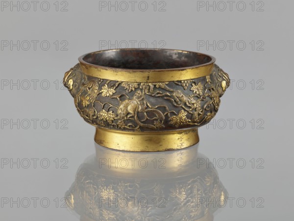Gui-type incense burner, Li, Daguan fl. late 16th–early 17th century? author signature, Chinese art, Chinese culture, Qing 1644–1911, zoomorphic motifs, gui vessels, plant ornament, ceremonial vessels, squirrels, vines, lions iconography, grapes, handles, ritual vessels, 18th/19th century, China, vessel, incense burner; height 6.5 cm, diameter 11.7 cm, weight 0.56 kg, A small metal bowl with a round foot and a flared rim sits centered on a reflective surface, showing a faint mirrored image below. The bowl has ornate raised decoration around its body featuring animal and plant motifs in a metallic gold tone against a darker brownish-gray background. The rim, foot, and some decorative elements are bright gold, while the interior and recessed areas are a darker, almost blackened brown. A pair of sculpted handles in gold protrude from opposite sides of the bowl, each shaped with a stylized animal head. The overall color palette includes gold, dark brown, gray, and black.