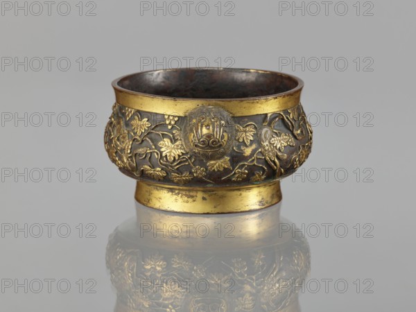 Gui-type incense burner, attributed to Li Daguan fl. late 16th–early 17th century? signed, noted; Chinese, Qing dynasty 1644–1911; bronze vessel with zoomorphic motifs—squirrels, lions—vines, grapes and plant ornament; gui vessel form with handles; ritual/ceremonial incense burner; 18th–19th century; height 6.5 cm, diameter 11.7 cm, weight 0.56 kg., A small ornate bowl sits against a plain gray background, with a reflective surface beneath showing a muted mirror image; the bowl has a rounded upper section and a narrower foot, both with bands of bright gold color, while the main body shows darker bronze and brown tones; the exterior is decorated with raised floral and vine motifs in gold relief and a few circular medallion-like decorations, and the interior appears dark with brown and black hues; overall the visible colors include gold, bronze, brown, black, and gray.