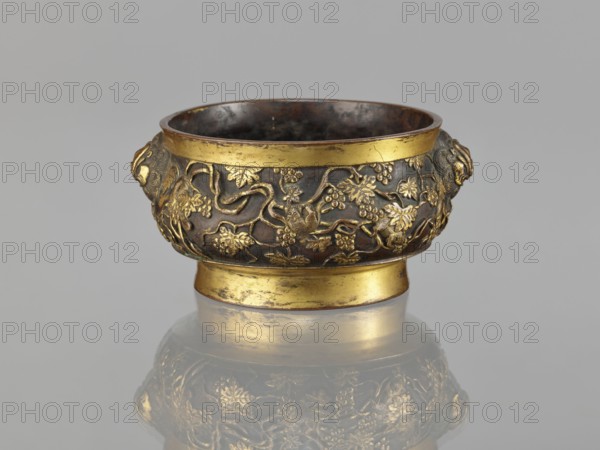 Gui-type incense burner, Li, Daguan fl. late 16th - early 17th century?,  foundryman author signature, Chinese art, Chinese culture, Qing 1644-1911, zoomorphic motifs, gui vessels, plant ornament, ceremonial vessels, squirrels, vines, lions iconography, grapes, handles, ritual vessels, 18th/19th century, China, vessel, incense burner, height 6.5 cm, diameter 11.7 cm, weight 0.56 kg, A small ornate bowl with a rounded body and a short pedestal base sits centered against a neutral gray background, displaying a dark metallic surface with areas of bright gold embellishment; the exterior features raised decorative vines and leaves in gold relief against a darker bronze backdrop, and there are sculpted lion head handles on opposite sides with gold detailing; the rim and the pedestal band are gilded, and the interior appears darker with a worn metallic look; the bowl casts a slightly blurred reflection beneath it.