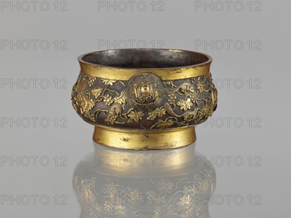Gui-type incense burner, Li, Daguan fl. late 16th–early 17th century? signature, Chinese art, Chinese culture, Qing 1644–1911, zoomorphic motifs, gui vessels, plant ornament, ceremonial vessel, squirrels, vines, lions iconography, grapes, handles, ritual vessel, 18th/19th century, China, vessel, incense burner; height 6.5 cm, diameter 11.7 cm, weight 0.56 kg, A small ornate bowl with a rounded body and a short pedestal base sits against a light gray background, showing metallic surfaces in gold and dark brown; the upper and lower rims are gold while the midsection is dark brown with raised gold decorative motifs depicting a dense pattern of leaves, vines, and animal figures, and a round medallion-like ornament on the front; the interior appears dark and slightly reflective, and the object casts a soft reflection on the surface beneath it.