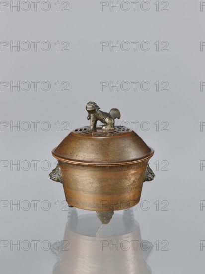 Censer with cover, Qing dynasty 1644–1911, 19th century, China. Censer with two handles in the shape of mascarons; lion fo iconography. Ritual vessel, inlaid decoration with gold veins. Height 12.5 cm, width 11.1 cm., An ornate lidded metal vessel with a warm bronze and darker brown patina sits centered on a reflective surface, featuring a domed lid topped by a small sculpted animal figure in a darker metallic tone, pierced decorative openings around the lid beneath the figure, a rounded body with subtle engraved patterns and horizontal banding, sculpted animal-head handles on the sides, three short ornate feet with cloud-like motifs, and a muted gray background; visible colors include bronze, brown, darker metallic gray, and pale gray in the reflection.