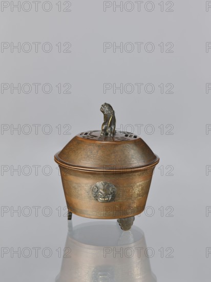 Censer with cover, Qing 1644–1911, Chinese, 19th century. Censer with two handles in the shape of mascarons; lion fo iconography; handles; ritual vessel. Decoration: inlay with gold vein. Materials: vessel censer. Dimensions: height 12.5 cm; width 11.1 cm., A small lidded metal vessel sits on a reflective surface, the vessel showing a warm bronze color with darker bronze or black accents; the lid has a perforated pattern and a small sculpted animal handle in dark bronze, and the body is decorated with engraved patterns and a round sculpted mask-like ornament on one side; the vessel stands on a group of short feet in dark bronze, and the reflection below shows muted bronze and gray tones against a pale gray background.