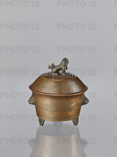 Censer with cover, Qing dynasty 1644–1911, 19th century, China. Censer with two handles in the shape of mascarons; lion fo iconography. Ritual vessel, censer; inlay with gold vein. Height 12.5 cm, width 11.1 cm., A bronze vessel with a domed lid and a stylized animal finial stands on a reflective surface; the vessel is primarily brown and bronze with darker greenish and blackish patina accents, a few decorative relief patterns around its body, a group of ornate handles shaped like animal heads on its sides, and three short, elaborately carved legs.