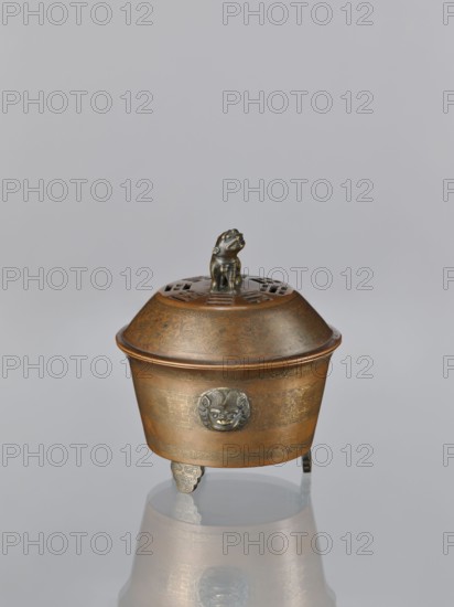 Censer with cover, Qing 1644–1911, Chinese, censer with two handles in the shape of mascarons lion/fo iconography, ritual vessel, inlay with gold vein decoration, 19th century, China, height 12.5 cm, width 11.1 cm, A metallic container with a domed lid sits against a neutral gray background, the container showing a warm bronze color with darker bronze and muted gold detailing, and a small sculpted figure in a darker metallic hue perched on top of the lid; the lid has pierced decorative openings and etched patterns, the body of the container displays engraved motifs and a raised circular mask-like ornament in a darker metallic tone, and the piece stands on a few short ornate feet that cast a subtle reflection on the glossy surface beneath it.