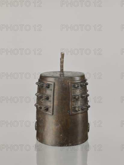 Bell linzhong, Chinese; Qing dynasty Daoguang, 1821–1850, dated 1825; idiophone percussion musical instrument used in religious rites; decorated with ideograms and meander ornament; height 29.0 cm, width 20.0 cm, depth 17.0 cm., A cylindrical metal object with a rounded top and a short vertical handle rising from its center is shown against a light gray background; the object appears in brown and dark bronze tones with areas of darker patina and slight greenish spots, and it has a few rectangular raised panels on its sides each studded with a few rounded rivet-like protuberances, with the base reflected faintly on the glossy surface below.