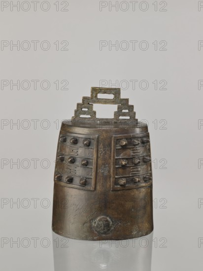 Bell linzhong, from Ignacy Jan Paderewski 1860–1941; Chinese; Qing dynasty 1644–1911, Daoguang period 1821–1850; 1825; bronze musical instrument idiophone/percussion; used in music and religious rites; decorated with ideograms and meander ornament; height 29.0 cm, width 20.0 cm, depth 17.0 cm., A metal bell-like object sits against a pale background, with a stepped rectangular handle on top showing a patterned surface and open rectangular cutouts; the body below is tapered and dark bronze in color with areas of lighter brown and greenish patina, and the upper section has a few raised bosses arranged in rows on panels separated by vertical bands; the lower section is smoother with a rounded decorative element near its base that shares the same bronze and greenish tones.