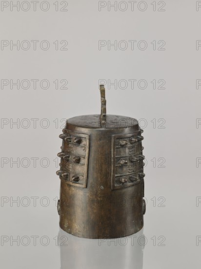 Bell linzhong, from Ignacy Jan Paderewski, Chinese, Qing dynasty Daoguang, 1821–1850, 1825, bronze percussion idiophone used in music and religious rites; decorated with ideograms and meander ornament. Dimensions: height 29.0 cm, width 20.0 cm, depth 17.0 cm., A cylindrical metal object with a rounded top and a vertical handle rising from its center sits against a plain light gray background, showing a patina of brown and dark bronze tones with areas of lighter greenish discoloration; the sides feature rectangular raised panels on opposite faces, each studded with a few rounded protrusions arranged in rows, and the base reflects faintly on the smooth surface beneath it.