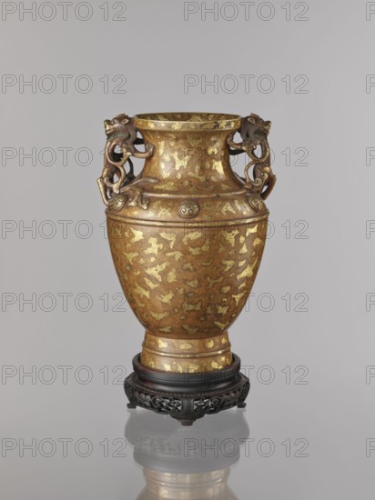 Vase on base., : Paderewski, Ignacy Jan 1860–1941. Chinese, Qing 1644–1911. Cast bronze vase with inlay of cold gold, dusting over lacquer; varnished bronze with full-plastic relief and engraved decoration. Decorated on the shoulders with six buttons with rosettes and an antidated signature; lion iconography. On a high foot. Round base with bulging side decorated with openwork plant ornament, on four short legs one missing. 17th/18th century, China. Vessel, vase: height 26 cm, diameter 16.5 cm; base: height 5.3 cm, diameter 13.1 cm., A gilded metal vase sits on a dark carved wooden stand; the vase has a warm golden brown surface with irregular gold patches and a rounded body that narrows at the neck and flares at the rim, and a pair of ornate handles shaped like stylized animal or dragon figures in a darker bronze tone curving from the neck to the shoulder.