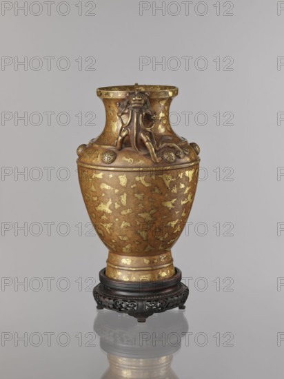 Vase on base. Chinese art; Chinese, Qing 1644–1911. Cast in bronze with cold-gold inlay, dusted over lacquer; varnished bronze with full-plastic relief and engraved decoration. On a high foot. Shoulders decorated with six buttons with rosettes. Round base with bulging side decorated with openwork plant ornament, on four short legs one missing. Lion iconography; antidated signatures. 17th–18th century, China. Vessel, vase: height 26 cm, diameter 16.5 cm; base: height 5.3 cm, diameter 13.1 cm., A metallic vase with a rounded body and flared neck sits on an ornate dark wooden stand, the vase showing a patinated bronze color with irregular gold-colored patches across its surface; the neck features sculpted decorative elements including a stylized animal or mythical creature relief and rounded knop-like details, all sharing the same bronze and gold tones, while the stand is carved with openwork patterns in a deep brown to black color and the background is a smooth pale gray.