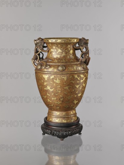 Vase on base. Chinese, Qing 1644–1911. 17th/18th century. Cast in bronze with cold-gold inlay, dusting over lacquer; varnished bronze, full-plastic relief and engraved decoration. On a high foot. Shoulders decorated with six knop-like buttons with rosettes. Round base with bulging side decorated with openwork plant ornament, on four short legs one missing. Lion iconography. Antidated signatures. Vessel; vase: height 26 cm, diameter 16.5 cm; base: height 5.3 cm, diameter 13.1 cm., A decorative vase with a rounded body and flared neck sits on an ornate carved stand; the vase shows a metallic brown surface with irregular gold patches and accents, and it has sculpted handles shaped like stylized animal heads and looping forms attached near the neck, with raised decorative bands and circular medallion-like elements around the shoulder; the stand is dark wood with intricate openwork carving, and the scene is set against a neutral light gray background with the vase and stand reflected faintl...