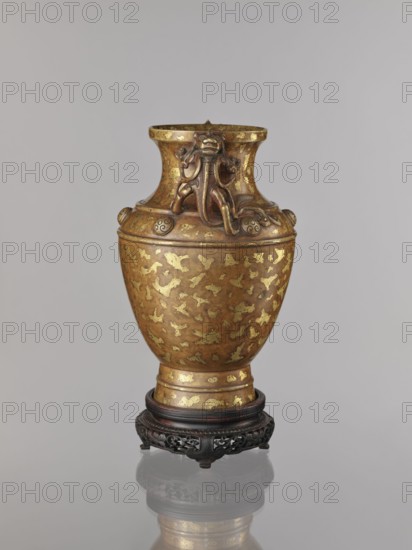 Vase on base. Chinese, Qing 1644–1911. 17th/18th century. Cast bronze with cold-gold inlay, dusting over lacquer; varnished bronze with full-plastic relief and engraved decoration. Decorated on the shoulders with six buttons with rosettes; lion iconography. Round base with bulging side decorated with openwork plant ornament, on four short legs one missing. Vessel vase: height 26 cm, diameter 16.5 cm. Base: height 5.3 cm, diameter 13.1 cm., A single ornate vase sits centered in the image on a carved wooden stand, showing a warm metallic surface with gold and brown tones and irregular gold leaf-like patches across its body; the vase has a flared neck with sculpted decorative elements resembling stylized animal or mythical motifs in a slightly darker bronze hue, and rounded ornamental bosses around the shoulder; the stand is dark brown to black with intricate openwork carving, and the whole object is reflected faintly on a glossy pale gray surface against a smooth light gray background.