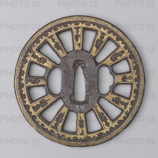 Crossguard tsuba, Shôami, and Migaki ; tsuba of the kuruma tsuba type, of the Buddhist Law Circle; oval seppa dai; on both sides decoration of irregular brass inlaid spots, and on the edge two stripes, the outer one filled with fine dots. Unsigned. 18th century Edo period, 1603–1868, Japan. Tsuba; diameter 8.5 cm, thickness 0.5 cm., A round, flat metal object with a central elongated oval opening and a few surrounding irregular cutouts, featuring radial openwork segments separated by raised bars; the surface shows areas of brown iron and patches of gold-toned inlay or gilding, with darker brown decorative cloud-like motifs on the gold sections and a textured band along the outer rim; the background is a uniform light gray.