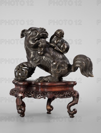Incense burner in the shape of lions on a base; creator, ; Chinese art; Chinese culture; Qing dynasty 1644–1911, 19th century; censers with covers; lion iconography; incense burner with a base; sculpture/ vessel/ censer; height 13.0 cm, width 8.0 cm, depth 20.0 cm., A sculpted animal-like figure with a curled tail and stylized mane sits on an ornately carved wooden stand; the figure appears metallic with a dark bronze color and subtle lighter highlights, while the stand shows reddish-brown and darker brown wood tones. The metal figure has rounded, bulbous forms on its back and a decorative spherical object near one paw, and the wooden base features intricate openwork carving along its apron and curved, cabriole-style legs. The background is a plain light gray.