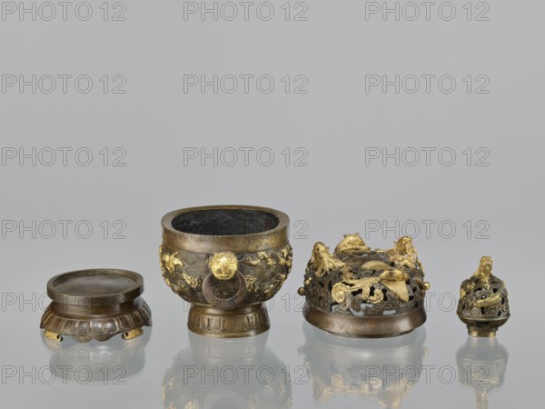 Censer with a cover on a base;, Qing period 18th century, China. Bronze vessel censer with high foot, openwork cover and handles; cover has removable knob. Decoration: dragons and ho-o phoenix birds on the cover; various animals on the belly including tigers, birds, fish; motifs of dragon with a pearl, clouds, sea waves, geometric patterns meander. Ritual/ceremonial vessel. Height 22.0 cm, width 21.0 cm, depth 17.7 cm., A group of small metal objects arranged in a row on a reflective surface against a neutral gray background: a low round container with a ridged base and a flat lid in dark bronze tones with some golden accents; a larger bowl-shaped vessel with a wide rim, ornate relief decoration and a dark interior, showing bronze and gilded areas; a domed, openwork cover richly decorated with sculpted creatures and floral forms in mixed bronze and bright gold; and a smaller ornate finial-like piece with openwork decoration and gilded detailing. The visible colors include bronze, dark ...