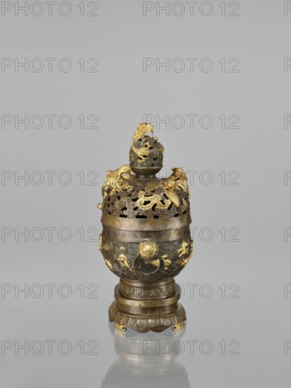 Censer with a cover on a base, Qing style, Chinese, 18th century. Bronze ceremonial vessel censer with high foot, openwork cover and handles; cover with removable knob. Decoration: dragons and hôô phoenix birds on the cover; various animals tigers, fish, birds and sea waves, clouds, dragon with a pearl, meander and geometric patterns on the body. Height 22.0 cm; width 21.0 cm; depth 17.7 cm., An ornate metal vessel stands against a neutral gray background, showing a combination of dark metallic and bright gold tones. The vessel has a rounded body on a tiered base with decorative fluted and banded details in dark metal with gold highlights. The upper section features an openwork lid with intricate pierced patterns and applied sculptural elements in gold, including leafy and animal-like forms. Gold ornamentation also appears on the body as raised figures and decorative motifs contrasting with the darker metal surface. A finial on top is similarly detailed with openwork and gold accents. ...