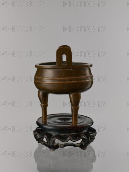 Ding incense burner on stand, Chinese, Chinese art, happiness symbol, lotuses, ceremonial vessel, Qing 1644–1911, bats iconography, one hundred antiquities baigu ornament, meander ornament, fo iconography, tiger symbol, ritual vessel. Decoration: lotus motifs, stylized hieroglyphs FU happiness. Round wooden turned stand on five legs. Sides of the stand are in the shape of a double lotus rosette lower part without the top. Upper base on foot separately. 19th century, China. Vessel, censer, incense burner: height 21 cm, diameter 15 cm; base: height 7.9 cm, width 18.4 cm., A bronze tripod vessel with a rounded bowl and a looped handle rising from its rim stands on three tapered legs atop a dark wooden circular stand with a carved openwork base; the vessel is brown with subtle engraved patterns and the stand is dark brown to nearly black, all set against a pale gray background.
