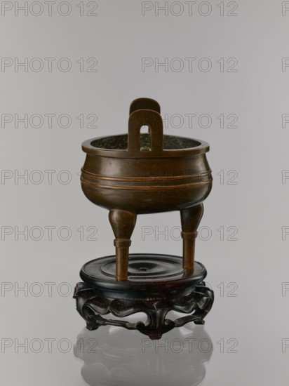 Ding incense burner on stand;, Chinese, Qing dynasty 1644–1911, 19th century. Bronze vessel censer incense burner. Decoration: lotus motifs, stylized hieroglyphs FU happiness, bats iconography, tiger symbol, fo iconographic, one hundred antiquities baigu ornament, meander ornament. Function: ritual/ceremonial vessel. Round wooden turned stand on five legs; sides shaped as double lotus rosette lower part without the top; separate upper base on foot. Dimensions: incense burner height 21 cm, diameter 15 cm; base height 7.9 cm, width 18.4 cm., A brown metal censers with a rounded body and a pair of vertical handles sits on a dark brown wooden stand with carved openwork legs; the censer has a polished surface with subtle engraved patterns and rests on three tapered legs above a circular top on the stand, and the visible colors include brown and dark brown against a light gray background.