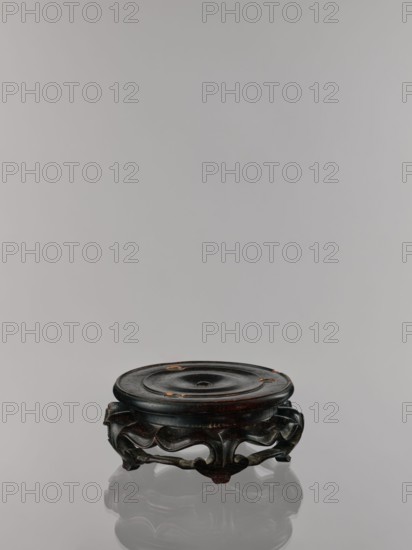Ding incense burner on stand, Chinese, Qing dynasty 1644–1911. Incense censer ritual vessel, 19th century. Round wooden turned stand on five legs; sides shaped like double lotus rosette lower part without the top; upper base on foot separate. Decoration: lotus motifs, stylized hieroglyph FU happiness; iconography includes bats, fo, tiger, lotuses, one hundred antiquities baigu, meander ornament. Dimensions: incense burner height 21 cm, diameter 15 cm; base height 7.9 cm, width 18.4 cm., A low, round wooden stand with a dark brown to almost black finish sits on a reflective surface, showing a subtle reflection beneath it; the top surface is slightly concave with a central shallow depression and a few small areas where the finish is worn to a lighter brown, while the carved base features flowing, curving cutouts and scrolled feet in the same dark brown hue with some lighter brown highlights.