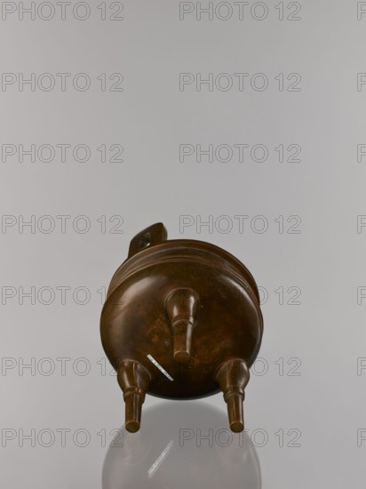 Ding incense burner on stand, maker, Chinese, Qing dynasty 1644–1911; 19th century. Bronze vessel censer/incense burner with decorations of lotuses, bats iconography, fo iconography, tiger symbol, meander and one hundred antiquities baigu ornament; stylized hieroglyph FU happiness. Round turned wooden stand on five legs; sides shaped as double lotus rosette lower part without the top; separate upper base on foot. Dimensions: burner height 21 cm, diameter 15 cm; base height 7.9 cm, width 18.4 cm., A brown rounded vessel with a handle on its upper side rests on three short cylindrical legs, showing a smooth polished surface with subtle darker brown variations and faint decorative incised lines; the background is a pale gray and the vessel casts a soft reflection below it.