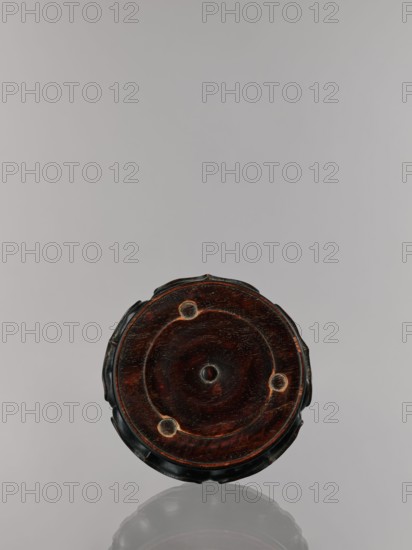 Ding incense burner on stand, Chinese, Qing 1644–1911, 19th century. Bronze ritual vessel and censer incense burner with decoration: lotus motifs, stylized hieroglyphs FU happiness, bats iconography, tiger symbol, one hundred antiquities baigu ornament, meander ornament, fo iconographic. Round wooden turned stand on five legs; sides shaped as a double lotus rosette lower part without the top; separate upper base on foot. Dimensions: incense burner height 21 cm, diameter 15 cm; base height 7.9 cm, width 18.4 cm., A round wooden object with a dark brown surface and lighter brown grain patterns sits against a pale gray background, showing a circular raised center and a faint concentric ring with three small circular indents around it; the edge appears black with scalloped contours and the object casts a subtle reflection below.