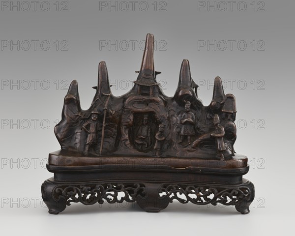 Brush stand on base, Chinese, Chinese art, mountains, painting implements, Qing 1644–1911, men, horses, scholars, Figure of a Hermit in a cave and other figures, 19th century, China, varia; height 21.0 cm, width 10.0 cm, depth 25.0 cm, A carved dark brown wooden sculptural scene sits on a carved base with scrolling openwork; the upper carving resembles a jagged mountain ridge with a central taller peak and several smaller peaks, and a group of carved human figures arranged along the front, some standing and some seated, with one figure holding a long staff and another seated in a small recessed niche; the surface shows polished highlights and darker recesses, with the overall colors being dark brown, deep reddish-brown, and small areas of black in shadow.