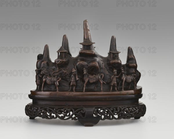 Brush stand on base, Chinese, Qing dynasty 1644–1911, 19th century. Materials: painting implements; subject: Figure of a hermit in a cave and other figures, men, scholars, horses, mountains. Dimensions: height 21.0 cm, width 10.0 cm, depth 25.0 cm. Function: brush stand., A dark brown carved wooden sculptural object resembling a stylized mountain range with pointed peaks and small pavilion-like structures, set on an oval base with an intricately carved openwork stand; the foreground features a group of small carved figures and animals positioned along the base of the mountain, all rendered in the same dark brown material with polished highlights and subtle variations in brown tones, against a neutral gray to light gray gradient background.