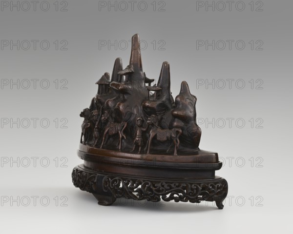 Brush stand on base, Chinese, Qing dynasty 1644–1911, 19th century. Materials: painting implements; subject: Figure of a Hermit in a cave and other figures, men, scholars, horses, mountains. Dimensions: height 21.0 cm, width 10.0 cm, depth 25.0 cm., A dark brown carved wooden sculpture sits on an ornate stand of similar dark brown wood; the carving depicts a group of mountain-like forms with smooth rounded peaks and ledges, small pavilion-like structures perched on some ledges, and a procession of human figures and animals arranged along a curved base. The figures and animals are carved in relief, with visible limbs and simple clothing or saddles, and a few raised platforms and rounded protrusions create depth against the mountain shapes. The stand features an openwork scrolling pattern and rests on short carved feet. The overall color palette is shades of dark brown with subtle variations from polished highlights to deeper recessed shadows, set against a neutral gray-to-white gradient...