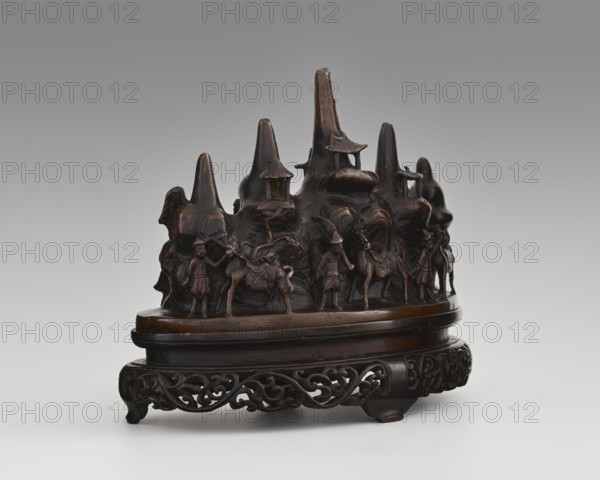 Brush stand on base, Chinese, Qing dynasty 1644–1911, 19th century. Materials and function: painting implements. Subject: Figure of a hermit in a cave and other figures — men, scholars, horses, mountains. Dimensions: height 21.0 cm, width 10.0 cm, depth 25.0 cm., A carved dark brown wooden sculpture depicting a mountain-like form with several pointed peaks and small pavilion-like structures, sitting on an oval base atop an intricately carved low stand; around the base of the peaks are a group of human figures and a few animals rendered in relief, some standing and some appearing to lead or accompany the animals, with detailed clothing and saddles; the stand and base show ornamental openwork and scrolling motifs; the visible colors are dark brown, lighter brown highlights on raised areas, and a neutral gray background.